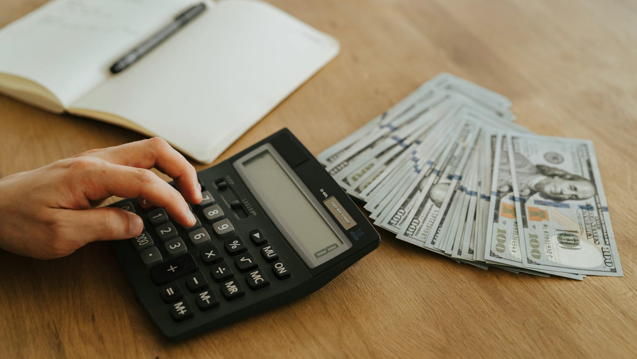 Close-up of a hand using a calculator with cash and a notebook on a wooden table.
